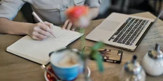 Henkilö kirjoittaa muistiinpanoja, pöydällä myös mm. tietokone