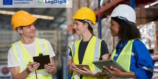 kolme ihmistä varastohallissa kypärät päässä ja suojaliivit päällä neuvottelemassa