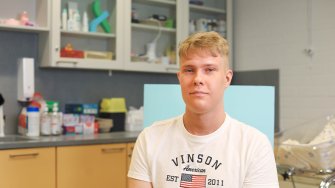 LAB-ammattikorkeakoulun terveydenhoitajaopiskelija Onni Laitinen istuu ja hymyilee kameralle. Kuvan taka-alalla näkyy hoitotyön luokan hyllyjä, joissa on erilaisia hoitotarvikkeita.