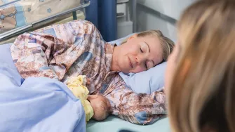 Nainen sairaalavuoteessa imettää vauvanukkea toisen naisen katsoessa vieressä