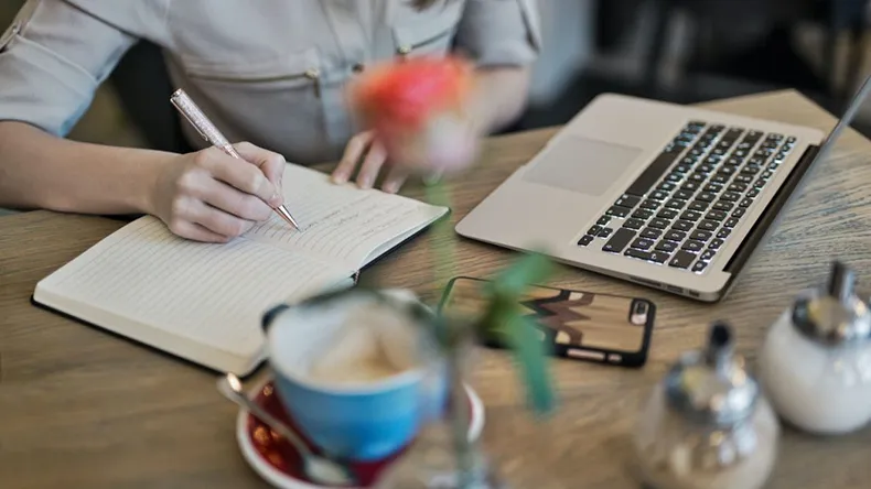 Henkilö kirjoittaa muistiinpanoja, pöydällä myös mm. tietokone