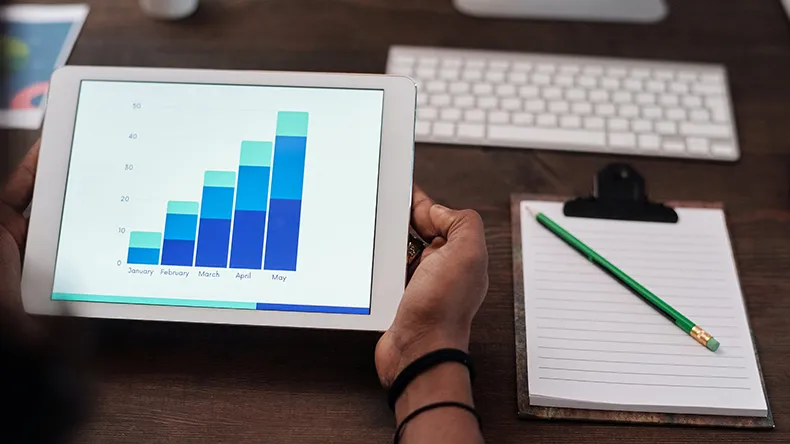 Ihmisellä kädessään tablettitietokone, vieressä muistiinpanot työpöydällä