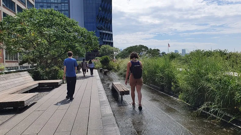 People walking on a sidewalk with water on one side. Vegetation and buildings in the background.