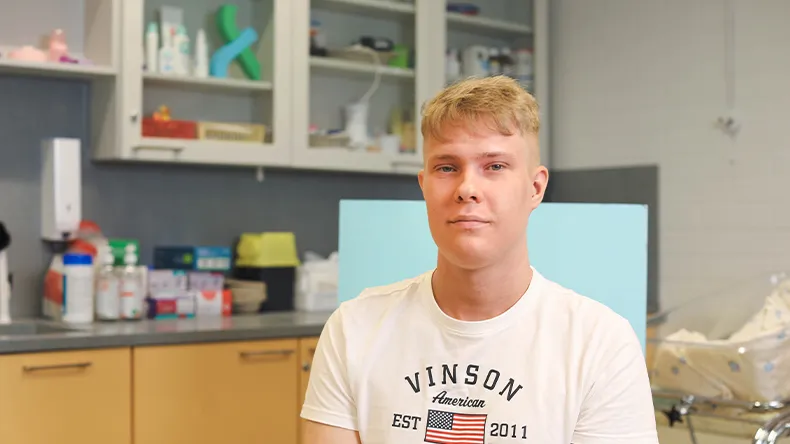 LAB-ammattikorkeakoulun terveydenhoitajaopiskelija Onni Laitinen istuu ja hymyilee kameralle. Kuvan taka-alalla näkyy hoitotyön luokan hyllyjä, joissa on erilaisia hoitotarvikkeita.