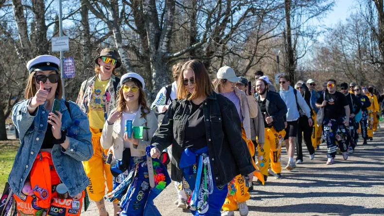 Kuvassa opiskelijoiden muodostama vappukulkue. Opiskelijoilla eri värisiä haalareita, etualan tyttö näyttää peukkua. 