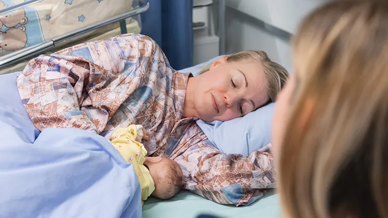 Nainen sairaalavuoteessa imettää vauvanukkea toisen naisen katsoessa vieressä