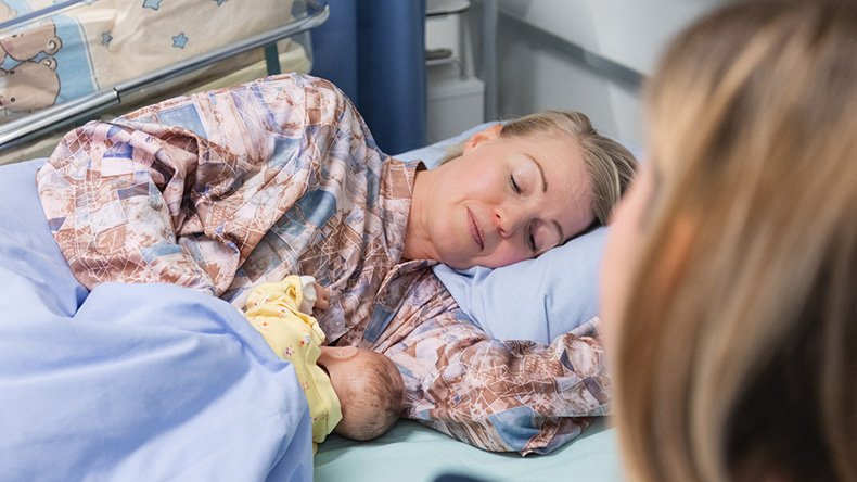 Nainen sairaalavuoteessa imettää vauvanukkea toisen naisen katsoessa vieressä